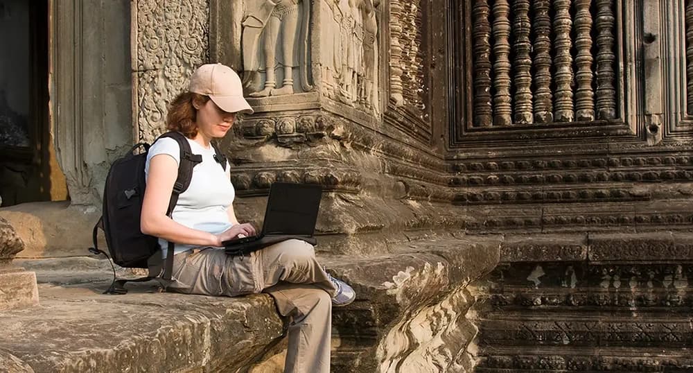 Woman in baseball cap sits on a stone ledge, working on her laptop