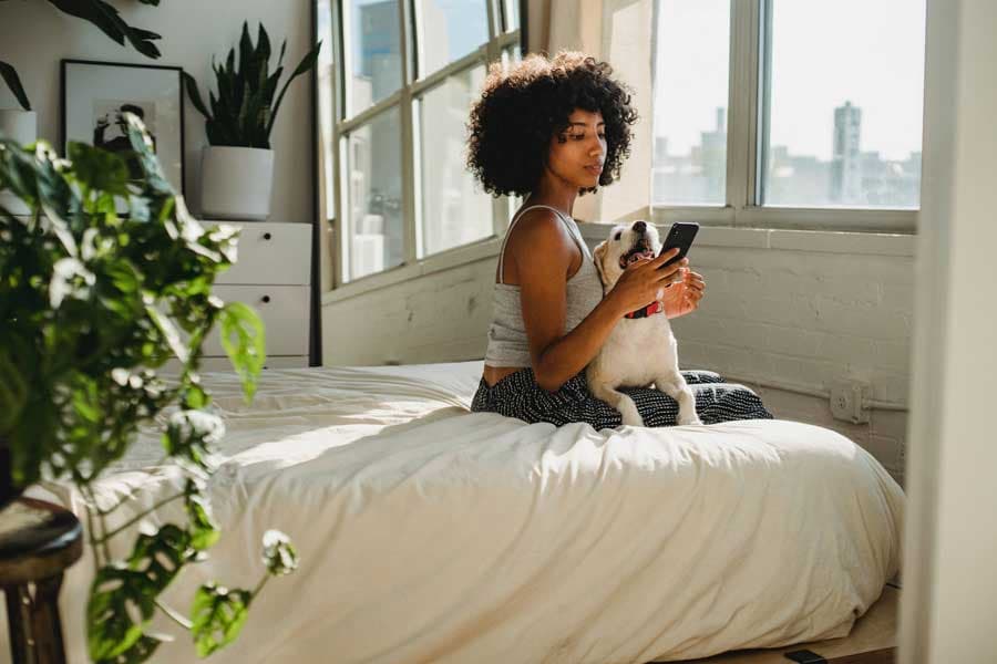 Woman sits with her tiny dog on a bed, looking at a cell phone