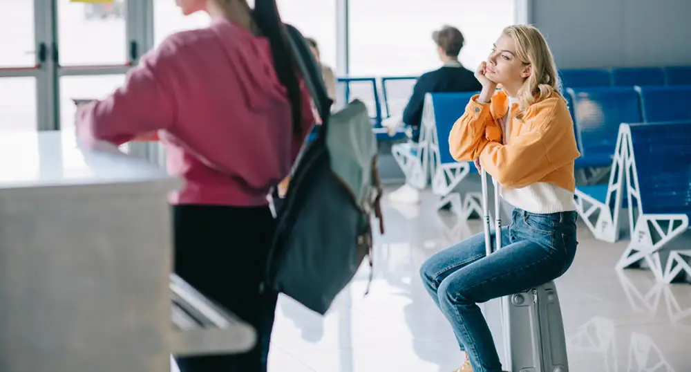 Woman sits on her suitcase while waiting in line at the airport